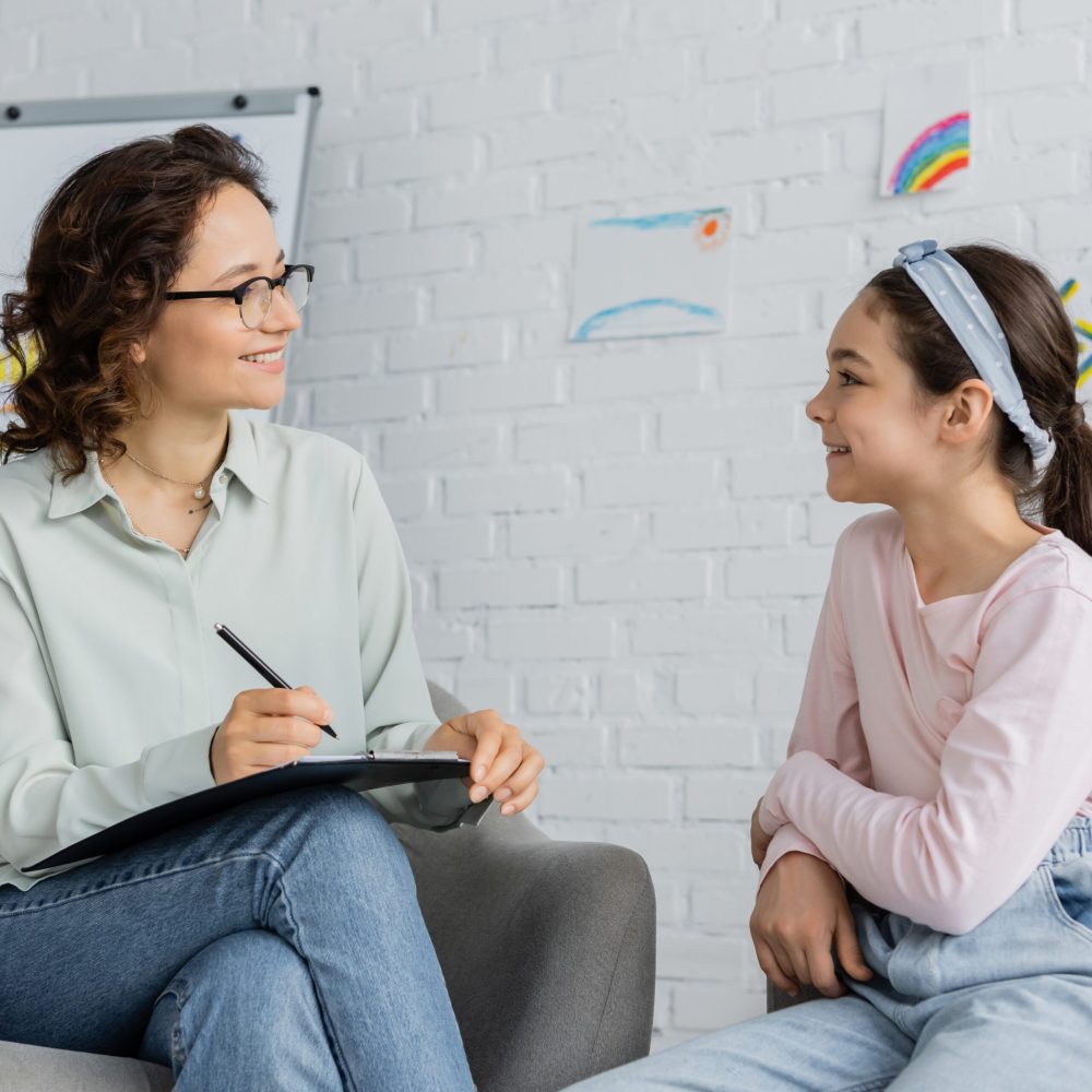 Smiling psychologist in eyeglasses holding clipboard and looking at preteen daughter in consulting